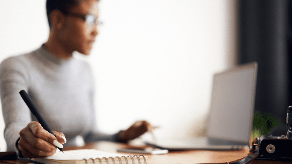 A dark-skinned woman works at her computer and writes in a notebook.